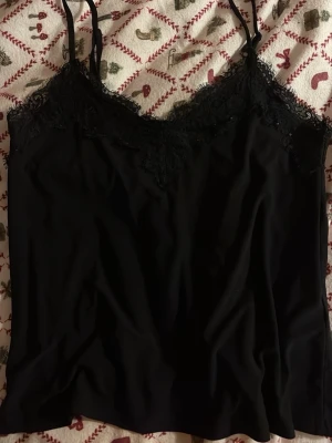 Svart linnetopp med spets från Amisu (new Yorker)  - Säljer en svart linnetopp från Amisu i storlek XS. Toppen har tunna, justerbara axelband och vacker spetsdetalj längs halsringningen. Materialet är mjukt och lätt, perfekt för att styla med jeans eller kjol. Tillverkad i Turkiet.