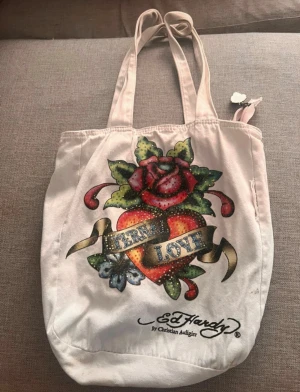 Ed Hardy shoppingväska med tryck - Säljer en vit shoppingväska från Ed Hardy med stort färgglatt tryck av hjärta, ros och texten 'Eternal Love' på framsidan. Väskan har långa handtag och dragkedja med tigerberlock. Materialet är bomull och designen är riktigt ikonisk. Se bilder för hål etc.💕