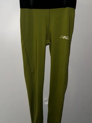 Olivgröna träningsleggings - Snygga olivgröna leggings med smal passform och elastisk midja. Materialet är stretchigt syntet, perfekt för träning. Diskret vitt tryck på ena låret ger en sportig touch. Passar dig som gillar att röra på dig och vill ha bekväma tights med stil.