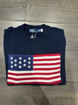 Mörkblå stickad tröja Polo Ralph Lauren - Snygg mörkblå stickad tröja från Polo Ralph Lauren med amerikansk flagga på bröstet. Tröjan har rund hals, ribbade muddar och RL-broderi i rött. Perfekt för dig som gillar klassisk och sportig stil.