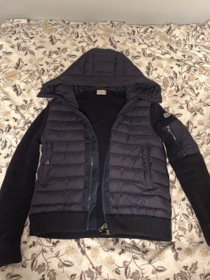 Navy blue Moncler cardigan  - Säljer en  Moncler cardigan med stickade ärmar och huva. Jackan har ribbade muddar, dragkedja framtill och fickor med både knapp och zip. Moncler-logga på ärmen och extra ficka med dragkedja på armen. Strorleken är 1 alltså S men passar även xs