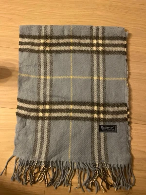Rutig blå ullhalsduk från Burberrys - Säljer en klassisk rutig halsduk från Burberrys i mjuk lammull. Färgen är ljusblå med inslag av grått, vitt och gult. Halsduken har fransar i kanterna och ett snyggt Burberrys-märke. Perfekt för att ge din outfit en cool touch.