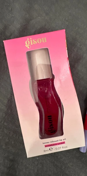 Gisou Honey Infused Lip Oil - Säljer en Gisou honey infused lip oil i färgen Strawberry Sorbet. Kommer i en snygg rosa förpackning och har en genomskinlig, kurvig tub med ljus lock. Innehåller 8 ml och ger läpparna en glansig, bärig finish med hint av honung.