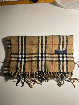 Beige rutig Burberry kashmirväst - Snygg väst från Burberry i klassiskt beige med rutigt mönster i svart, vitt och rött. Västen är tillverkad i 100% kashmir och har fransar längs nederkanten. Perfekt för dig som gillar lyxiga och tidlösa plagg med ikonisk design.