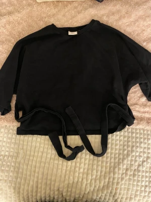 Svart omlottopp med knyt från Nelly - Svart omlottopp från Nelly med långa band för knytning i midjan. Toppen har en loose passform och är tillverkad i mjuk bomull. Rund halsringning och korta ärmar ger en chill vibe. Perfekt att styla med jeans eller kjol för en enkel look.