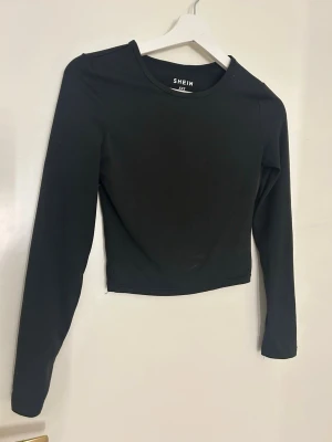 Svart långärmad topp från SHEIN - En stilren svart långärmad topp från SHEIN i storlek XXS. Toppen har rund halsringning och är croppad i modellen, vilket ger en modern och enkel look. Perfekt att matcha med jeans eller kjol för en clean vibe.
