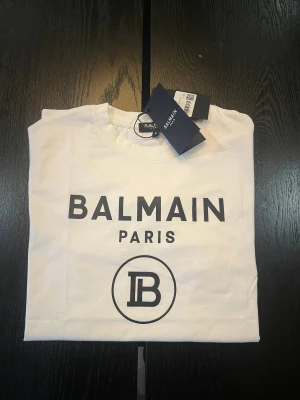 Vit Balmain Paris t-shirt i bomull - Snygg vit t-shirt från Balmain Paris med svart tryck framtill och rund hals. Klassisk passform och tillverkad i mjuk bomull. Perfekt för dig som gillar stilrena designer och vill ha ett statement-plagg med lyxig känsla. Storlek S.