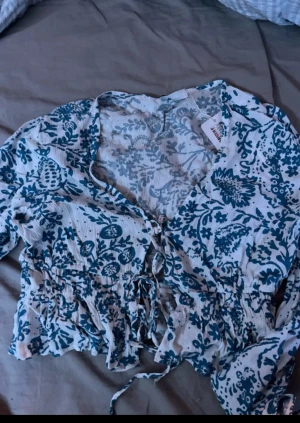 Vit blommig blus med snörning - Säljer en vit blus från FB Sister  med blått blommigt mönster. Blusen har lång ärm, v-ringad hals och snörning framtill. Croppad passform med volangdetaljer vid midjan och ärmslut. Perfekt för vår och sommar, superfin till jeans eller kjol.