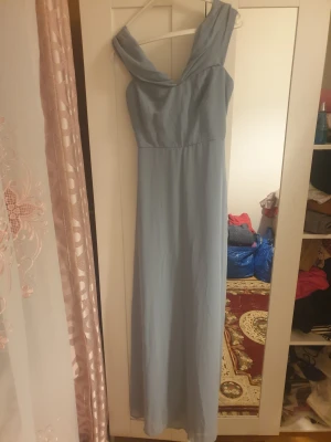 Ljusblå långklänning från Nily Eve - Säljer en elegant ljusblå långklänning från Nily Eve i storlek 36. Klänningen har en vacker omlottkrage och är ärmlös med en mjuk, flytande form. Materialet är lätt och känns som polyester. Dragkedja i sidan för enkel påklädning.