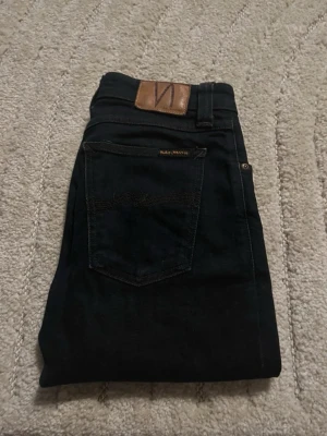  Nudie Jeans - Säljer ett par svarta jeans från Nudie Jeans med klassisk femficksdesign och läderpatch bak i midjan. Jeansen är slim fit och är tillverkade i ett slitstarkt bomullsmaterial. Perfekta för dig som gillar stilrena och tidlösa jeans.