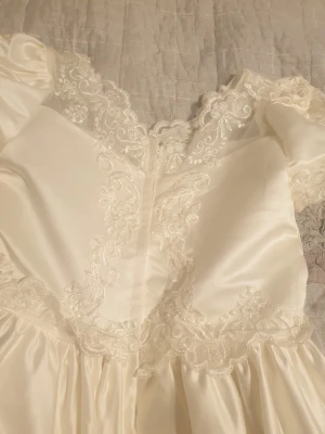 Vit långklänning med spetsdetaljer - Elegant vit långklänning med puffärmar och vacker spets längs halsringning, midja och nederkant. Klänningen har broderade detaljer och ett skirt, glansigt tyg som ger en lyxig känsla. Perfekt för speciella tillfällen och har en klassisk, romantisk look.