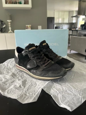 Philippe Model Trainers - Säljer ett par svarta Philippe Model Trainers med snygga detaljer i mocka och skinn. Skorna har snörning, rund tå och platt sula. Ovandelen är en mix av textil och skinn, med en diskret logga på sidan och en beige kant vid sulan. Perfekta för dig som gillar stilrena sneakers.