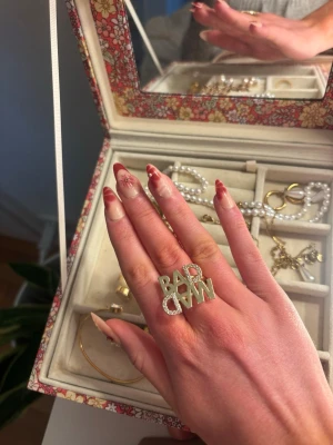Ring från Ioaku - Säljer en statementring från Ioaku med texten MAD BAD i guld och glittriga stenar. Ringen är stor och syns verkligen, perfekt för dig som vill sticka ut. Den är justerbar i storlek och har en cool design med stenar runt bokstäverna.