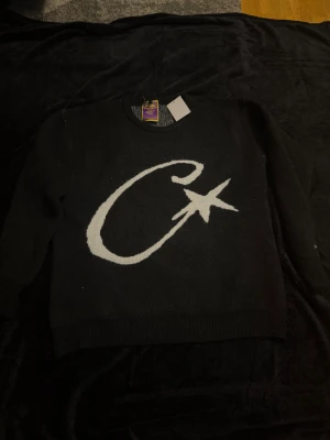 Corteiz sweater  - Cool svart stickad tröja från corteiz i storlek M med en stor vit C och stjärna på framsidan. Tröjan har rund hals, långa ärmar och ribbade muddar. Perfekt för dig som gillar grafiska detaljer och vill ha en unik look.