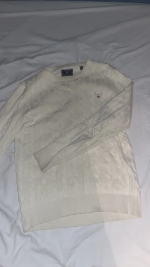 Vit kabelstickad tröja från GANT - Kabelstickad vit tröja från GANT i premium bomull. Klassisk rund halsringning, ribbade muddar och diskret GANT-logo broderad på bröstet. Perfekt för dig som gillar stilrena och tidlösa plagg.