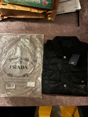 Svart overshirt jacka från Prada XL - Svart Prada overshirt i re-nylon med klassisk krage, silverfärgade knappar och fickor framtill. Snygg detalj med Prada-logga på bröstfickan. Perfekt för dig som gillar stilrena och exklusiva plagg med modern touch.