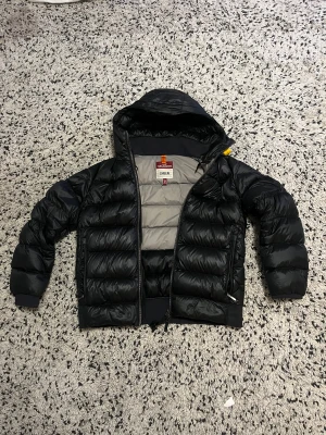 Parajumpers Pharell PufferJacka - Säljer en svart Parajumpers Pharell dunjacka i storlek M. Jackan har huva, dragkedja och quiltad design med grått foder. Materialet är glansigt och känns lätt men ändå varmt. Perfekt för kalla dagar och har snygga detaljer som orange hängare och märkning på insidan. Inga defekter alls på varan och orginal pris ligger om kring 6500 kr (620€)
