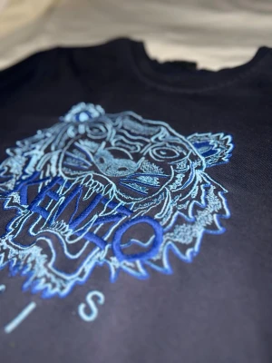 Mörk blå Kenzo sweatshirt med blå tiger - Mörkblå sweatshirt från Kenzo med ikonisk blått tigerbroderi och texten 'Kenzo Paris' på bröstet. Tröjan har rund hals, lång ärm och normal passform men är storlek M fast som också passar S. Materialet är mjuk bomull som känns skön mot huden. Perfekt för dig som gillar statement-plagg och snygga detaljer.