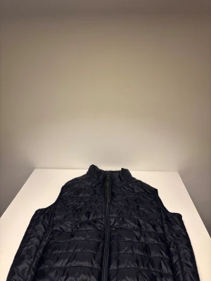 Mörkblå quiltad dunväst  - Mörklblå dunväst med quiltad design och hög krage. Västen har en hel dragkedja framtill och är tillverkad i ett glansigt syntetmaterial. Perfekt att slänga över en hoodie för extra värme. Enkel och stilren look som funkar till många outfits.