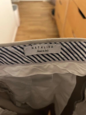 Beige kostymbyxor från Natalino - Chinos Natalino  Beiga chinos med hög midja från Natalino, tillverkade i Italien. Klassisk design med justerbara side adjusters, ”tungt” tyg och knappförsedda bakfickor. Liknande berg & Berg eller casatlantic.  Mått:  Innerbenslängd: 76cm Benöppning: 17,5cm Midja: 40,5cm Ytterbenslängd: 104 cm Front rise: 29,5cm
