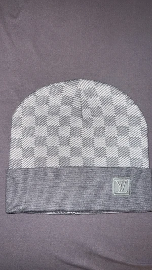 Grå rutig mössa Louis Vuitton - Snygg grå mössa från Louis Vuitton med klassiskt rutigt mönster och uppvikt kant. LV-logga broderad framtill. Materialet är mjukt och stickat, perfekt för kyliga dagar. Stilren design som lyfter din look.