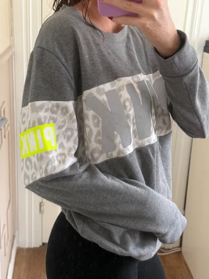 Grå sweatshirt från PINK med leopardmönster - Fin och skön grå sweatshirt från PINK med bredt vitt band över bröstet och ärmarna, dekorerat med grått leopardmönster och stor logga som är i reflex. Neongul PINK-detalj på ärmen. Rund hals och lång ärm, loose fit. 