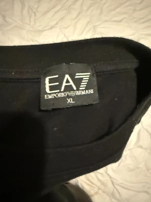 Svart EA7 Emporio Armani t-shirt XL - Svart t-shirt från EA7 Emporio Armani i storlek XL. Snyggt tryck i guld på bröstet med EA7-loggan. Klassisk rund hals och korta ärmar. Mjuk och skön bomullskvalitet som känns riktigt nice mot huden.
