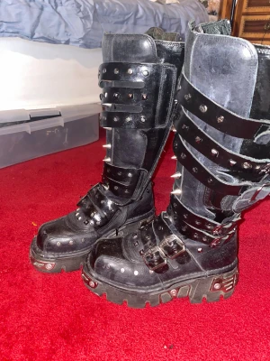 New Rock svarta boots med nitar - rejäla New Rocks stövlar i skinn med plattformsula, massor av metalldetaljer, nitar/spikes och spännen. höga skaft med dragkedja på insidan. de är äldre, går ej att köpa på hemsidan längre. stl 38, så för små för mig, om du har ett par i stl 39 är jag villig att byta. 