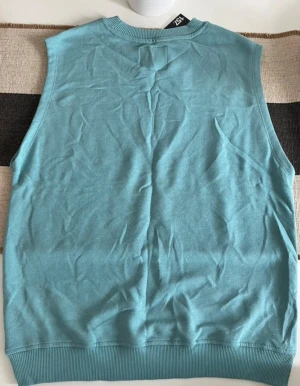 Turkos stickad väst från 157 - En stilren turkos stickad väst från 157 med ribbade kanter vid hals och nederkant. Västen har en enkel, rak passform och är perfekt för att lägga till en färgklick i din outfit. Mjuk och skön känsla tack vare det stickade materialet.