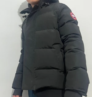 Svart pufferjacka från Canada Goose - Svart pufferjacka från Canada Goose med ikonisk röd och vit patch på ärmen. Jackan har hög krage med snörning, stora quiltade sektioner och är fylld med dun och fjädrar. Perfekt för kalla dagar och har en clean, modern look. Kommer med original förpackning. Pris kan diskuteras.