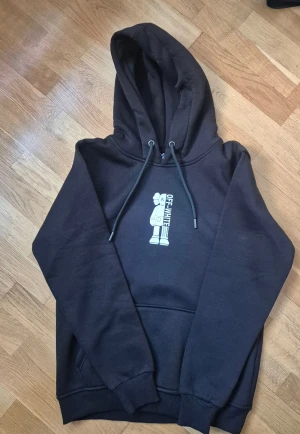 Svart Off-White hoodie med tryck - Svart hoodie från Off-White med stor huva och snörning. Framtill finns ett vitt tryck med Off-White-figur och på ryggen ett stort, färgglatt grafiskt tryck med olika motiv. Tröjan har ribbade muddar och en klassisk känguruficka. Perfekt för dig som gillar streetwear och coola prints.