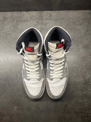 Nike Air Jordan 1 High Grey/White - Nike Air Jordan 1 High i grått och vitt med svarta detaljer. Ovandel i skinn och mocka, klassisk snörning och svart yttersula. Röd Nike Air-logga på plösen och ikonisk vinge på sidan. Perfekta för dig som gillar streetstyle och sneakers med retrovibe.