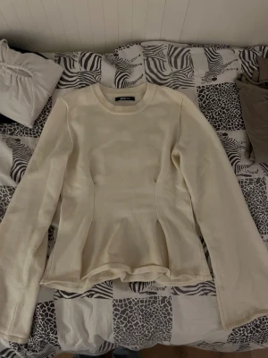 Krämvit peplumtop från Gina Tricot - Säljer en krämvit långärmad peplumtop från Gina Tricot. Toppen har rund halsringning, markerad midja och volang nedtill som ger en snygg siluett. Materialet är mjukt och stretchigt, perfekt för dig som gillar stilrena och feminina plagg. Har en liten sminkfläck vid kragen som förmodligen går bort i tvätten men annars använd fåtal gånger