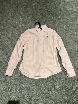 Rosa randig skjorta från Ralph Lauren - Snygg rosa och vit randig skjorta från Ralph Lauren i slim fit-modell. Skjortan har klassisk krage, långa ärmar och knappar framtill. Tillverkad i 100% bomull och pryds av den ikoniska broderade polospelaren på bröstet. Perfekt för dig som gillar stilrena och fräscha plagg.