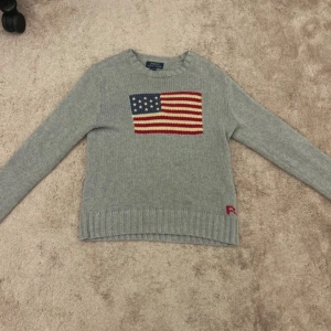 Grå stickad tröja Polo Ralph Lauren - Grå stickad tröja från Polo Ralph Lauren i 100% bomull. Framsidan har en stor amerikansk flagga i rött, vitt och blått som broderad detalj. Tröjan har rund hals, ribbade muddar och långärmad passform. Perfekt för dig som gillar klassisk och chill stil.