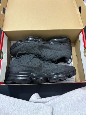 Nike Air Vapormax 2023 Svarta sneakers - Nike Air Vapormax 2023 sneakers i helsvart med stickad ovandel och synlig luftbubbelsula. Snygg, modern design med diskret Nike-logga på sidan och Vapormax-detalj på plösen. Skorna har snörning och en rund tå, perfekt för dig som gillar techiga och sportiga vibes.