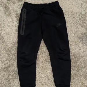 Svarta mjukisbyxor från Nike, storlek 137-147 - Svarta mjukisbyxor från Nike med snyggt tryckt logga på benet. Byxorna har snörning i midjan och en lång dragkedja på sidan för extra stil. Materialet är mjukt och bekvämt, perfekt för chill eller träning. Passformen är normal och benen är avsmalnande nedtill.