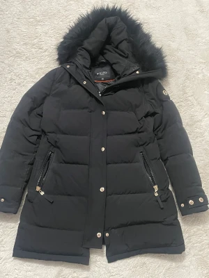 Svart pufferjacka från Hollies - Svart pufferjacka från Hollies med fluffig huva och guldfärgade detaljer. Jackan har långa ärmar, två fickor med dragkedja och snygga knappar vid ärmslut och framtill. Perfekt för kalla dagar med sin quiltade design och stilrena look.