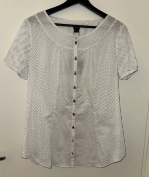 Vit prickig skjorta från Esprit - Snygg vit skjorta från Esprit med bruna prickar och korta ärmar. Modellen har rundad hals, knappar framtill och två dekorativa knappar på ärmsluten. Skjortan är tillverkad i mjuk bomull och har en loose passform som ger en chill vibe.