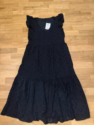 Svart långklänning med volang - Svart långklänning från VILA med broderade blommor och volang nedtill. Klänningen har v-ringning och korta volangärmar som ger en romantisk vibe. Tillverkad i mjuk bomull som känns skön mot huden. Perfekt för dig som gillar detaljer och en feminin siluett. Helt ny med prislapp, aldrig använd.