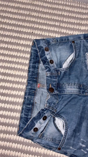 Blå jeans från Linde Industries - Säljer ett par blå jeans från Linde Industries med klassisk femficksdesign och slitna detaljer på framsidan. Jeansen har raka ben, knappgylf och snygga kontrastsömmar på bakfickan. Perfekt för dig som gillar en avslappnad stil. Model:slim fit 