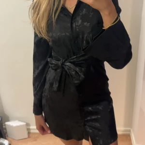 Svart omlottklänning med rosett - Snygg svart kortklänning med subtilt blommönster och omlott-design. Klänningen har långa ärmar, knytband i midjan som bildar en rosett. Materialet har en lätt glansig finish som ger en lyxig känsla. Använd en gång.