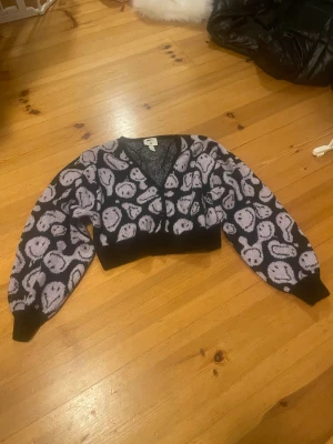 Lila kofta Smiley x H&M XS - Säljer en croppad lila och svart kofta från Smiley x H&M i storlek XS. Koftan har ett lekfullt smiley-mönster över hela plagget, v-ringad hals och knappar framtill. Materialet är stickat och ärmarna är långa med svarta muddar.