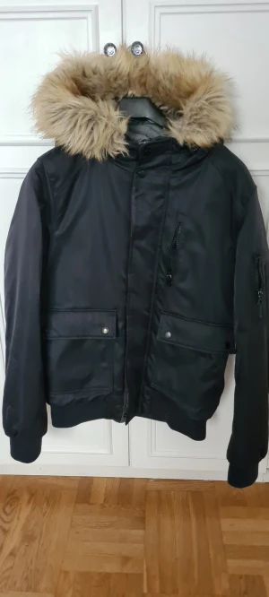 Svart bomberjacka med fuskpäls - Svart bomberjacka med stor beige fuskpäls på huvan. Jackan har flera fickor med knappar och dragkedjor, samt ribbade muddar vid ärmslut och nederkant. Perfekt för kalla dagar och har en snygg, urban look. Den är i storlek S
