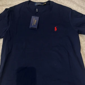 Mörkblå t-shirt från Polo Ralph Lauren - Snygg mörkblå t-shirt från Polo Ralph Lauren med klassisk röd logobrodering på bröstet. T-shirten har rund hals och korta ärmar, tillverkad i mjuk bomull för skön komfort. Perfekt för dig som gillar stilrena och tidlösa plagg.