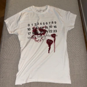 Maison Martin Margiela vit t-shirt - Säljer en riktigt snygg och eftertraktad T-shirt från Maison Martin Margiela i storlek S. T-shirten är i nära nyskick och använd mycket sparsamt.  Den ikoniska Margiela-designen med sifferprint på bröstet och det unika röd-abstrakta motivet gör den till ett statement-plagg som sticker ut utan att bli överdrivet. Passar perfekt både till vardags och för att lyfta en mer dressad outfit. Högkvalitativ bomull, skön passform. Perfekt för dig som gillar designerplagg med edge. Längd: 64 cm Bröst: 44cm