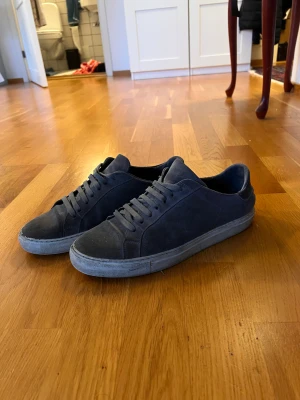Mörkgrå sneakers från Axel Arigato - Snygga mörkgrå sneakers från Axel Arigato i mocka med klassisk låg profil och rund tå. Skorna har platt sula och snörning framtill. Perfekta för dig som gillar stilrena och minimalistiska sneakers med premiumkänsla. Använda ett tag men inga större slitningar!