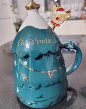 Julmugg med lock och sked - Charmig julmugg i form av en gran med lock, dekorerad med guldiga detaljer och medföljande sked med renmotiv. Muggen är i nyskick och levereras i originalförpackning – perfekt som julklapp eller till vinterns varma drycker.