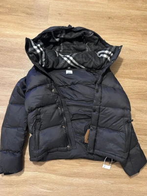 Svart pufferjacka från Burberry - Svart pufferjacka från Burberry med avtagbara ärmar, så du kan bära den som väst. Jackan har huva med rutigt foder, dragkedja och knappar framtill samt två fickor med dragkedja. Patch med Burberry London England på ena ärmen. Perfekt för kalla dagar.