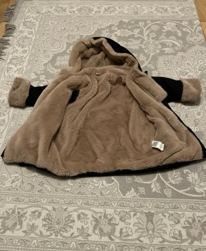 Svart teddyjacka med huva och beige foder - Säljer en svart teddyjacka med mysigt beige foder och huva. Jackan har dragkedja framtill, två fickor och uppvikta ärmslut med beige teddy. Perfekt för dig barn som vill ha en varm och chill look under kalla dagar. Passar 5 åringar 